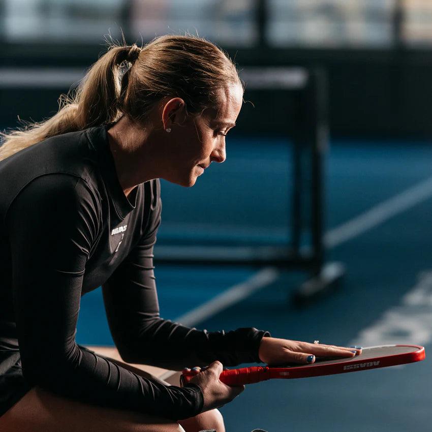 Frau in Sportkleidung sitzt mit Tennisschläger in der Hand auf einer Tennisplatzbank.