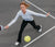 Frau in weißem Langarmshirt und schwarzer Hose spielt Pickleball und schlägt gelben Ball auf grauem Spielfeld.
